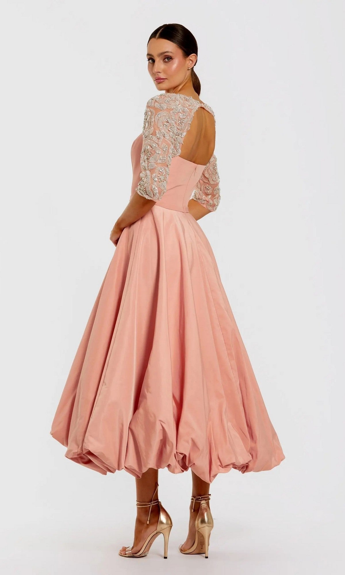 A woman models the Mac Duggal 2320 semi-formal midi party dress featuring a peach hue, flared skirt, embellished mesh sleeves, open back, and metallic high-heeled sandals against a plain background.